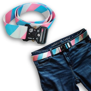 May include: A blue, pink, and white striped belt with a black buckle that says "RAWR" on it. The belt is shown on a pair of blue jeans with the words "AMERICAN EAGLE" on the back pocket.