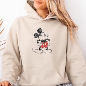May include: A beige hoodie featuring a black and red Mickey Mouse graphic. The graphic depicts Mickey Mouse with his arms crossed, wearing red shorts. The hoodie has a front pocket and a drawstring hood. The person is wearing blue jeans.