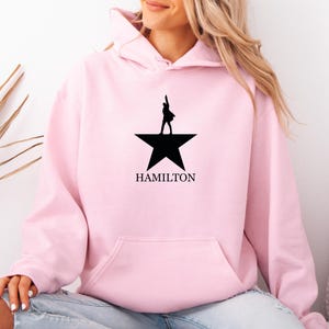 Peut inclure: Un sweat à capuche rose clair avec un motif noir représentant une personne sur une étoile et le mot "HAMILTON" en dessous. Le sweat à capuche a une poche avant et une capuche. La personne porte un jean bleu déchiré.