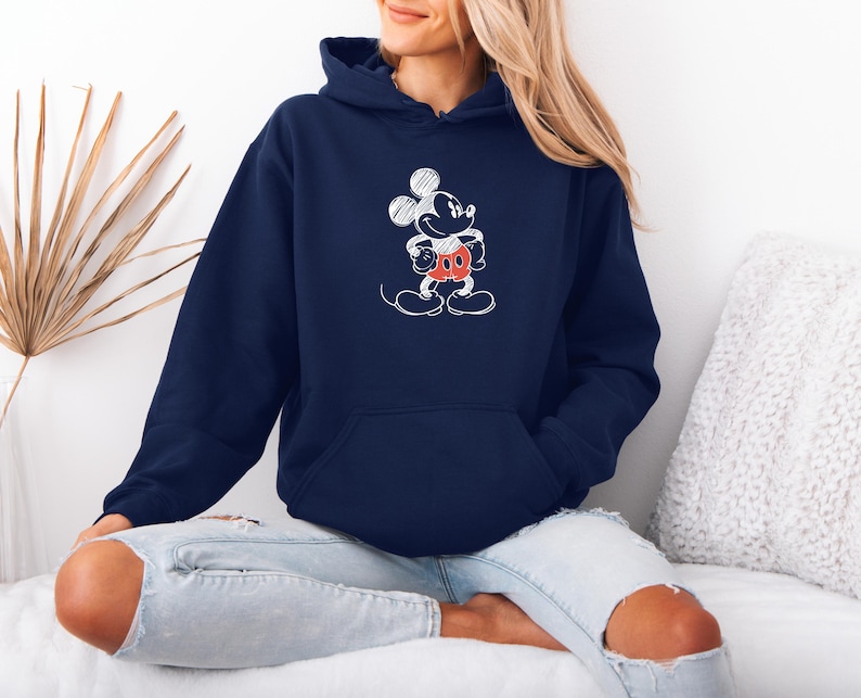 May include: Navy blue hoodie featuring a white sketch of Mickey Mouse. The cartoon character has a red and white outfit. The hoodie has a front pocket and a drawstring hood. The sweatshirt is made of a soft material.
