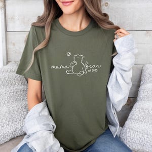 Pu&ograve; includere: Una maglietta verde con un contorno bianco di un orso e il testo "mama bear est 2025".