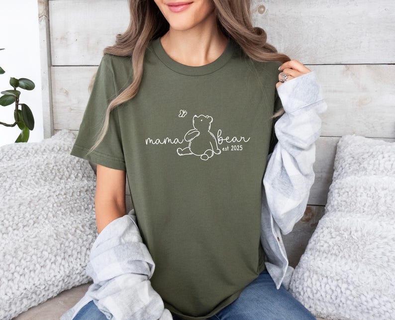Pu&ograve; includere: Una maglietta verde con un contorno bianco di un orso e il testo "mama bear est 2025".