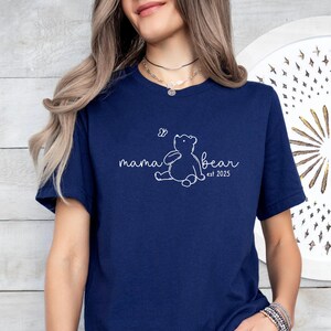 Pu&ograve; includere: Maglietta blu navy con un contorno bianco di un orso e una farfalla, e il testo "mama bear est 2023".