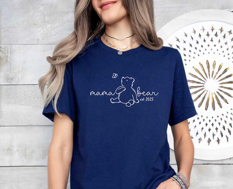 Pu&ograve; includere: Maglietta blu navy con un contorno bianco di un orso e una farfalla, e il testo "mama bear est 2023".