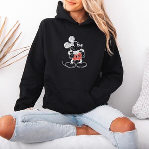 May include: Black hooded sweatshirt featuring a white sketch of Mickey Mouse with red shorts and a black face. The sweatshirt has a front pocket and a drawstring hood. The design is a classic cartoon character.