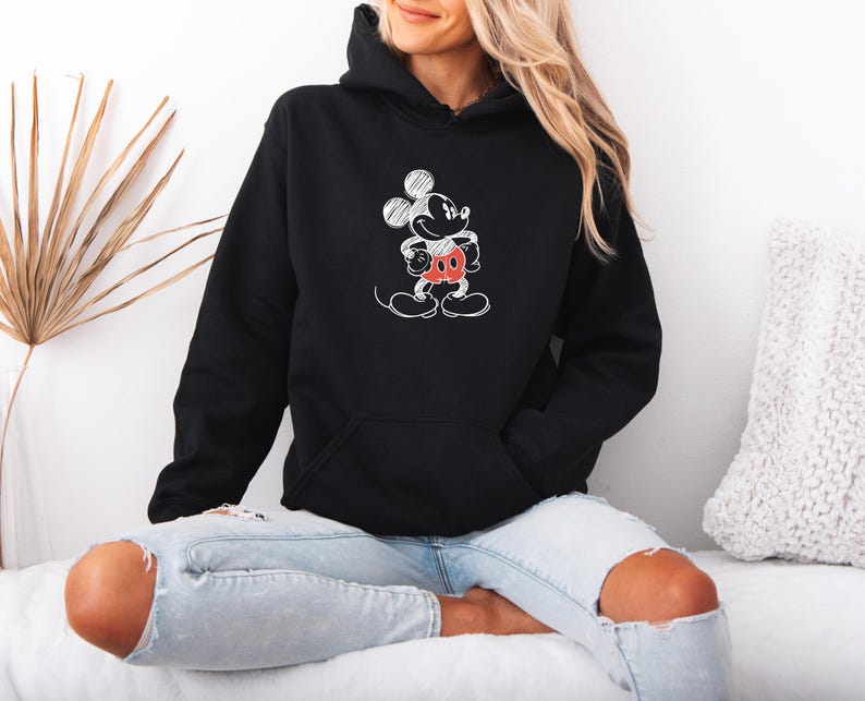 May include: Black hooded sweatshirt featuring a white sketch of Mickey Mouse with red shorts and a black face. The sweatshirt has a front pocket and a drawstring hood. The design is a classic cartoon character.