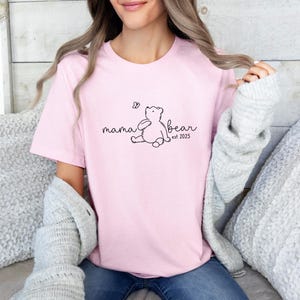 Pu&ograve; includere: Maglietta rosa con un contorno nero di un orso e il testo "mama bear est 2025".