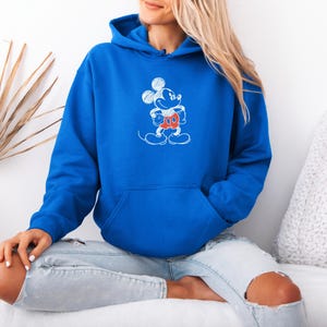 May include: Royal blue hoodie with a white and red Mickey Mouse sketch design. The hooded sweatshirt has a front pocket and a drawstring hood. The graphic features Mickey Mouse in a standing pose. The hoodie is made of a soft material.