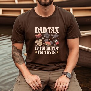 May include: A brown t-shirt with the text "DAD TAX" above cartoon images of Mickey Mouse and other characters. The text "IF I'M BUYIN' I'M TRYIN'" is below the images. The shirt is worn by a person.