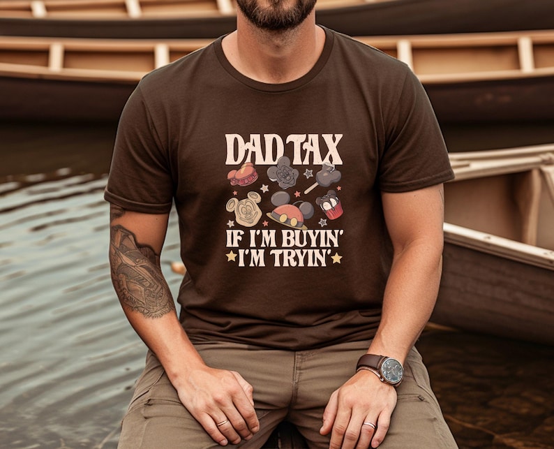 May include: A brown t-shirt with the text "DAD TAX" above cartoon images of Mickey Mouse and other characters. The text "IF I'M BUYIN' I'M TRYIN'" is below the images. The shirt is worn by a person.