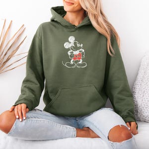 May include: Olive green hoodie featuring a white sketch of Mickey Mouse with red shorts. The hoodie has a front pocket and a drawstring hood. The design is a classic cartoon character. The model is wearing light blue jeans.