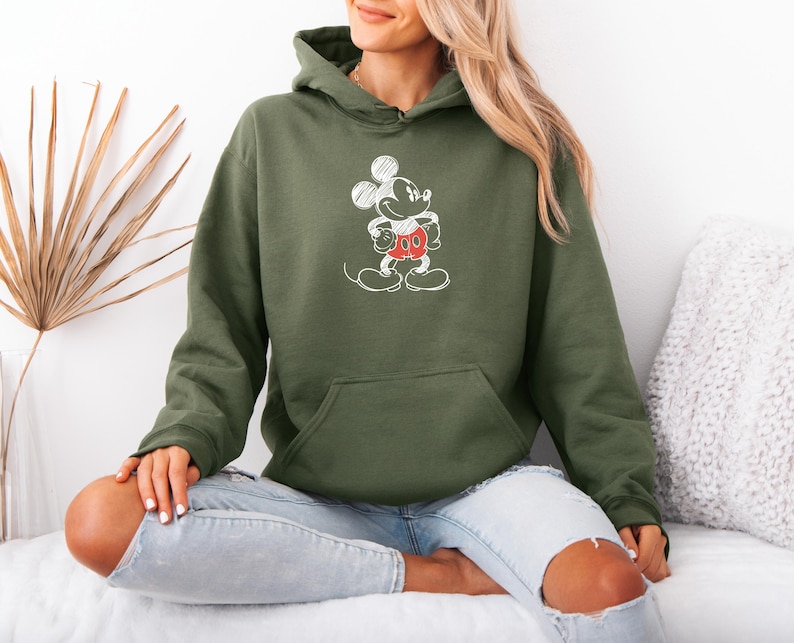 May include: Olive green hoodie featuring a white sketch of Mickey Mouse with red shorts. The hoodie has a front pocket and a drawstring hood. The design is a classic cartoon character. The model is wearing light blue jeans.