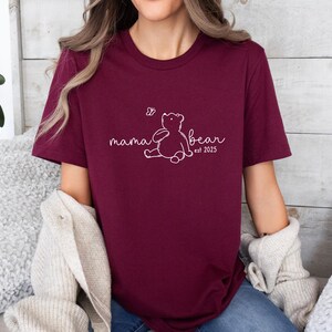 Pu&ograve; includere: Una maglietta color borgogna con un contorno bianco di un orso e il testo "mama bear est 2025".