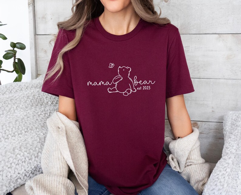 Pu&ograve; includere: Una maglietta color borgogna con un contorno bianco di un orso e il testo "mama bear est 2025".