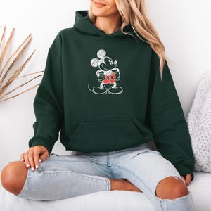 May include: Dark green hoodie featuring a white sketch of Mickey Mouse with red shorts. The hoodie has a front pocket and a hood. The person is wearing light blue jeans.