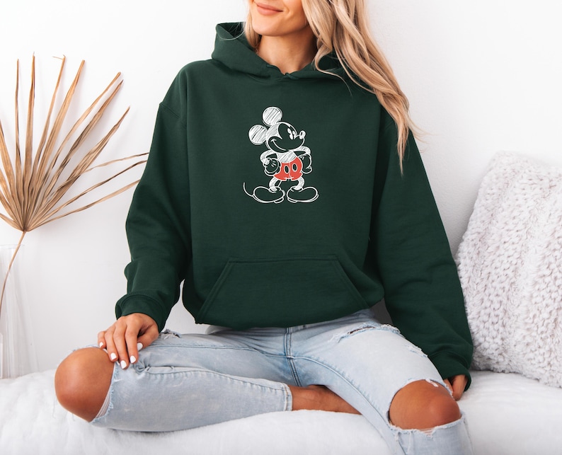 May include: Dark green hoodie featuring a white sketch of Mickey Mouse with red shorts. The hoodie has a front pocket and a hood. The person is wearing light blue jeans.