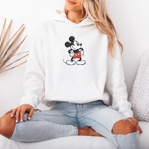 May include: White hooded sweatshirt featuring a black and red sketch of Mickey Mouse. The cartoon character is in a standing pose with his hands on his hips. The sweatshirt has a front pocket and is worn with light blue jeans.