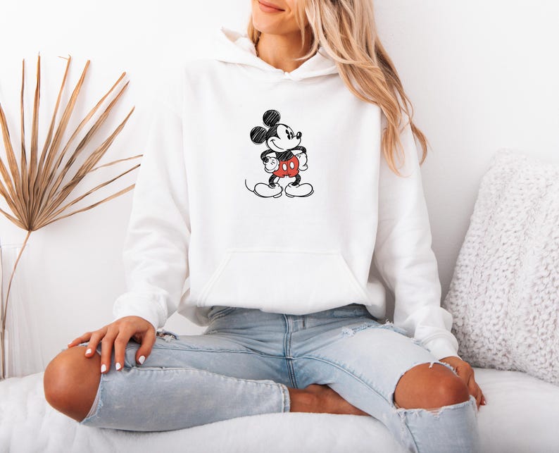 May include: White hooded sweatshirt featuring a black and red sketch of Mickey Mouse. The cartoon character is in a standing pose with his hands on his hips. The sweatshirt has a front pocket and is worn with light blue jeans.