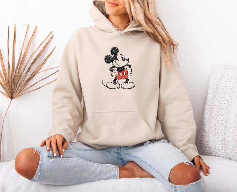 May include: A beige hoodie featuring a black and red Mickey Mouse graphic. The graphic depicts Mickey Mouse with his arms crossed, wearing red shorts. The hoodie has a front pocket and a drawstring hood. The person is wearing blue jeans.