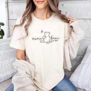 Pu&ograve; includere: Una maglietta bianca con una grafica nera di un orso e il testo "mama bear est. 2023".