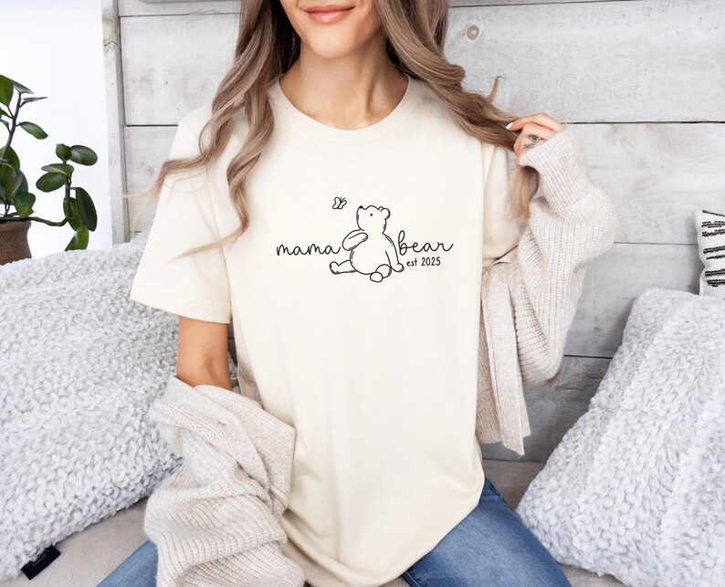Pu&ograve; includere: Una maglietta bianca con una grafica nera di un orso e il testo "mama bear est. 2023".