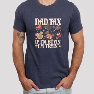 May include: Navy blue t-shirt with the text "DAD TAX" and the phrase "IF I'M BUYIN' I'M TRYIN'". The shirt features cartoon graphics of a Mickey Mouse head, a cupcake, and other themed designs. The shirt is a crew neck.