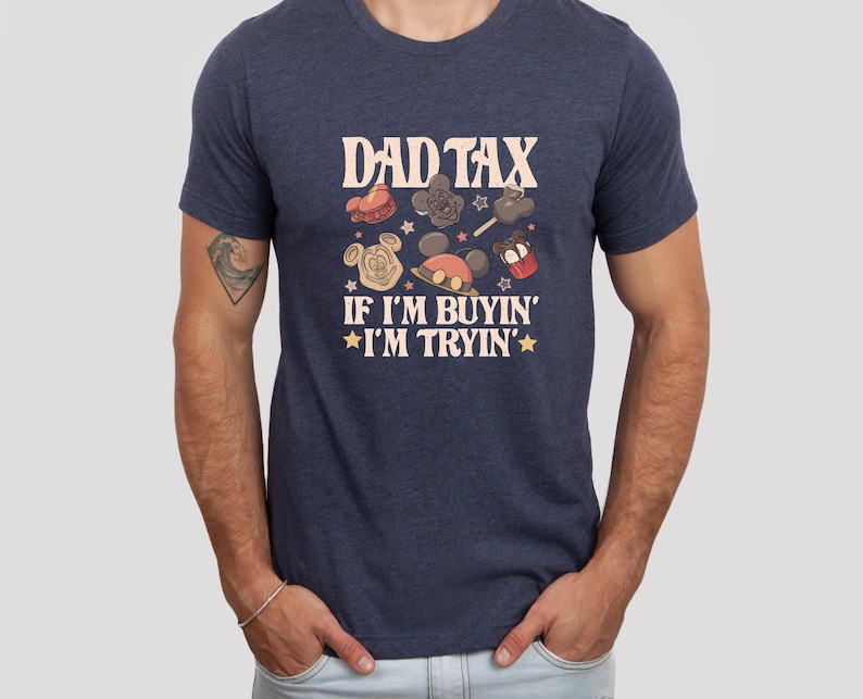 May include: Navy blue t-shirt with the text "DAD TAX" and the phrase "IF I'M BUYIN' I'M TRYIN'". The shirt features cartoon graphics of a Mickey Mouse head, a cupcake, and other themed designs. The shirt is a crew neck.