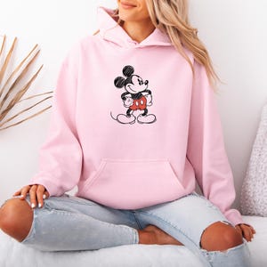 May include: A light pink hoodie featuring a black and red Mickey Mouse graphic. The hoodie has a front pocket and a drawstring hood. The graphic is a simple, sketched design of Mickey Mouse. The hoodie is made of a soft material.
