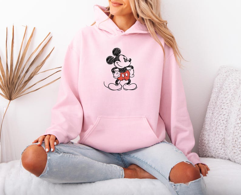 May include: A light pink hoodie featuring a black and red Mickey Mouse graphic. The hoodie has a front pocket and a drawstring hood. The graphic is a simple, sketched design of Mickey Mouse. The hoodie is made of a soft material.