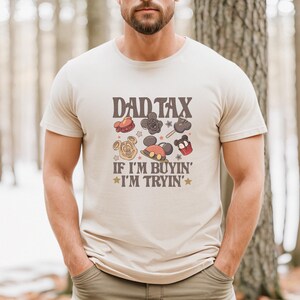 May include: Beige t-shirt with the text "DAD TAX" above cartoon images of theme park treats. Below the text is "IF I'M BUYIN' I'M TRYIN'" in a retro font. The shirt is worn by a person with a beard.