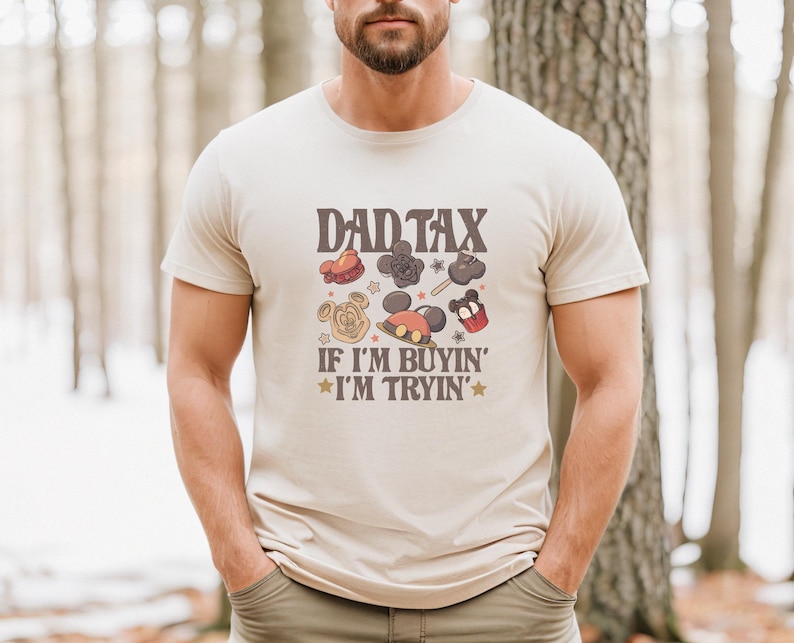 May include: Beige t-shirt with the text "DAD TAX" above cartoon images of theme park treats. Below the text is "IF I'M BUYIN' I'M TRYIN'" in a retro font. The shirt is worn by a person with a beard.