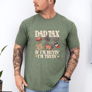 May include: Green t-shirt with the text "DAD TAX" above cartoon images of treats and Mickey Mouse-themed items. Below the text is "IF I'M BUYIN' I'M TRYIN'" in a retro font. The shirt is worn by a person.