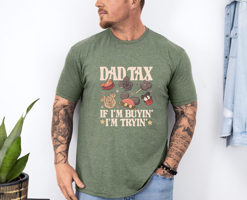 May include: Green t-shirt with the text "DAD TAX" above cartoon images of treats and Mickey Mouse-themed items. Below the text is "IF I'M BUYIN' I'M TRYIN'" in a retro font. The shirt is worn by a person.