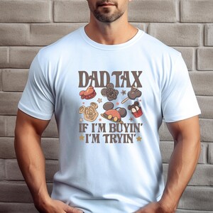 May include: Light blue t-shirt with the text "DAD TAX" above cartoon images of treats and the phrase "IF I'M BUYIN' I'M TRYIN'". The shirt features a crew neck and short sleeves.