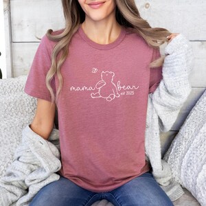 Pu&ograve; includere: Una maglietta rosa con un contorno bianco di un orso e il testo "mama bear est. 2025".