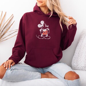 May include: Burgundy hoodie with a white sketch of Mickey Mouse on the front. The hoodie has a kangaroo pocket and a drawstring hood. The graphic features Mickey's face, hands, and shorts in red.