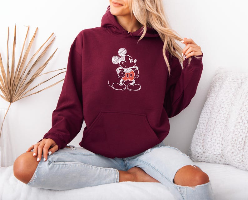 May include: Burgundy hoodie with a white sketch of Mickey Mouse on the front. The hoodie has a kangaroo pocket and a drawstring hood. The graphic features Mickey's face, hands, and shorts in red.