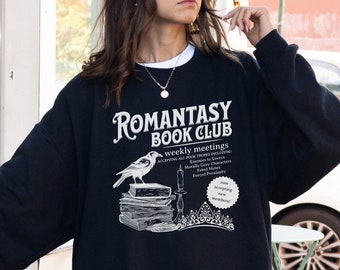 May include: A black sweatshirt with a white graphic print that says "Romantasy Book Club" with a bird, books, and a crown. The sweatshirt is being worn by a person.