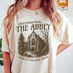 May include: Cream-colored t-shirt with a graphic that reads "Greetings from The Abbey, Home of the Sidhe-Seers." The graphic features a detailed illustration of a gothic church building, trees, and a path. The shirt is a casual, oversized fit.