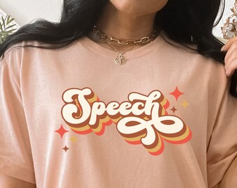 Può includere: T-shirt rosa chiaro con la scritta "Speech" in un font retrò. Il testo è stratificato con bianco, arancione e marrone. Piccole grafiche a forma di stella sono su entrambi i lati della parola.