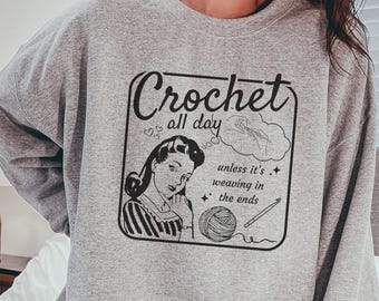 May include: A heather gray crewneck sweatshirt featuring a black graphic print of a person with the text Crochet all day unless it's weaving in the ends, accompanied by illustrations of a ball of yarn, a crochet hook, and small stars.
