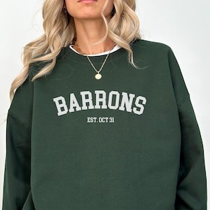 May include: A dark green sweatshirt with the word "BARRONS" in white block letters across the chest, with "EST. OCT 31" below. The sweatshirt is layered over a white shirt. The model is wearing black shorts.