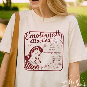 Könnte beinhalten: Cremefarbenes T-Shirt mit einem weinroten Aufdruck. Der Aufdruck lautet "Emotionally attached to my sourdough starter" und zeigt eine Illustration im Vintage-Stil einer Frau und Backutensilien.