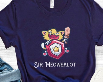 May include: Navy blue t-shirt featuring a cartoon cat character holding a sword and shield with a crown. The cat is smiling and the shield has a crown on it. The text "Sir Meowsalot" is printed below the graphic. The shirt is paired with blue jeans and white sneakers.