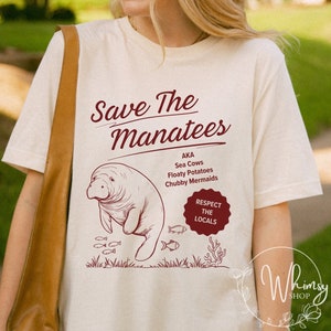 Puede incluir: Una camiseta beige con un gráfico marrón de un manatí con el texto "Save The Manatees" y "AKA Sea Cows Floaty Potatoes Chubby Mermaids" y un círculo rojo con el texto "Respect The Locals".
