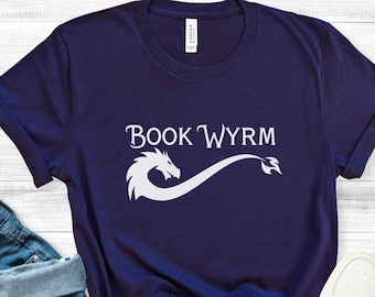 May include: A navy blue t-shirt with a white dragon design and the text "BOOK WYRMM" printed on the front. The shirt is laid flat on a white wooden surface.