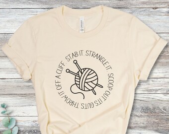 May include: A beige t-shirt with a black circle graphic. The graphic features a ball of yarn with knitting needles and the text "It's got its guts throw it off a cliff stab it strangle it scoop it out".