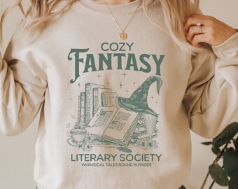 Może przedstawiać: Beżowa bluza z nadrukiem przedstawiającym kapelusz czarownicy, książki i filiżankę herbaty. Tekst na bluzie brzmi "Cozy Fantasy Literary Society Whimsical Tales Bound in Pages".