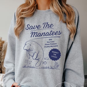 Puede incluir: Sudadera gris con un gráfico azul de un manatí con el texto "Save The Manatees" y "Respect The Locals".