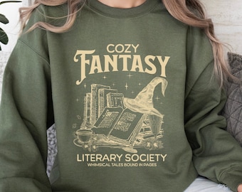 Może przedstawiać: Zielona bluza z napisem "Cozy Fantasy Literary Society Whimsical Tales Bound in Pages" i grafiką stosu książek z czarodziejskim kapeluszem na górze.